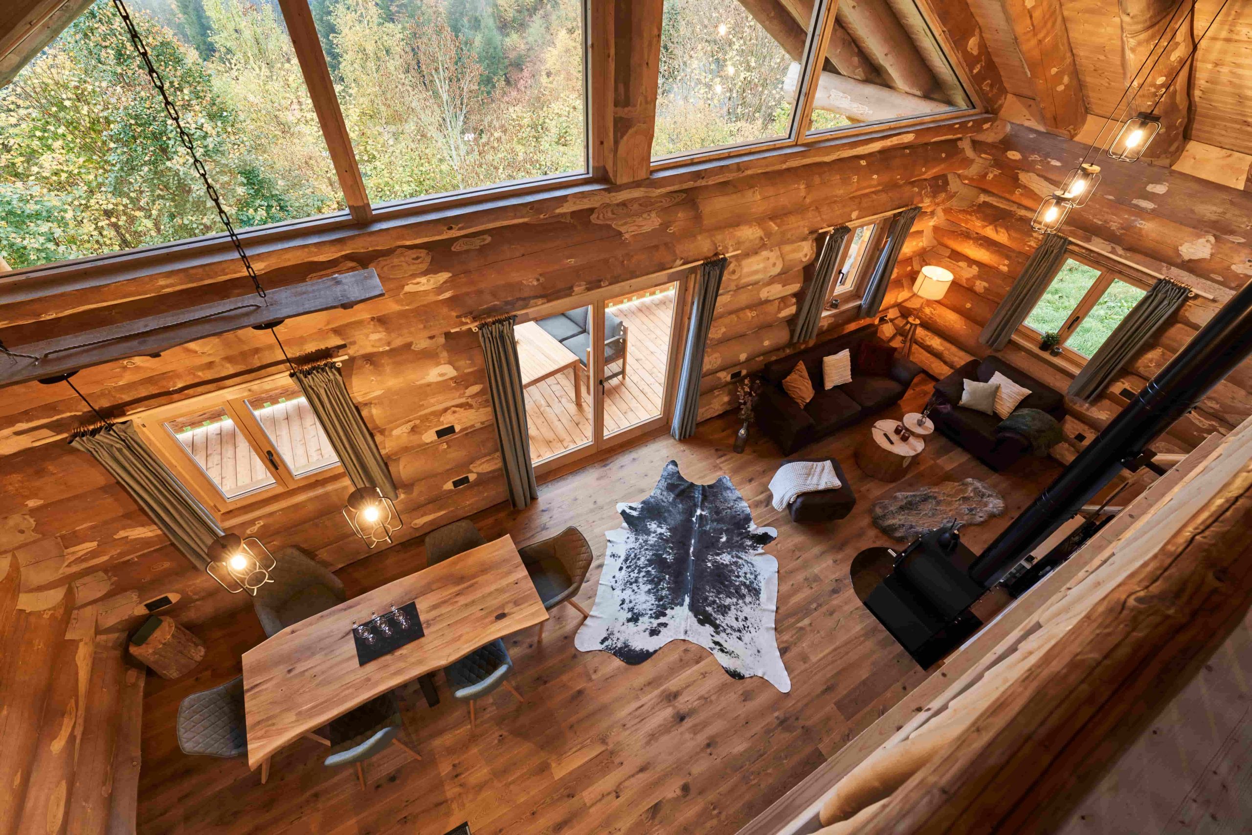 Beeindruckende Ansicht von der Galerie des Chalet Wipfelblick auf den gesamten Wohn- und Essbereich, den Kuhfell-Teppich und die großen Panoramafenster mit Blick ins Grüne