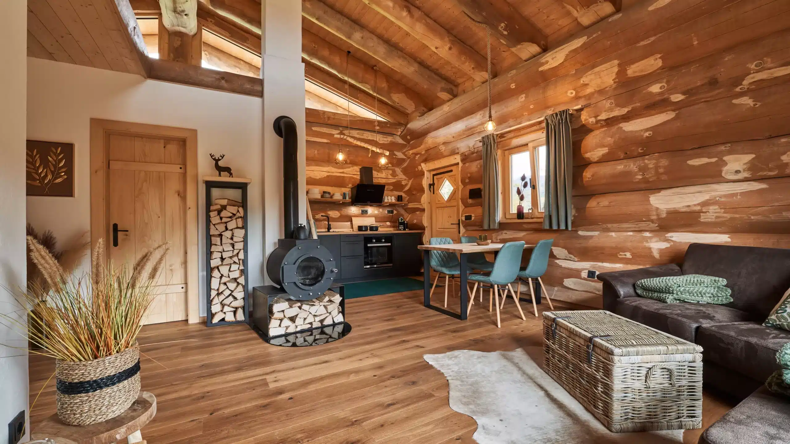 Moderner Wohnbereich in Chalet Feldstein mit schwarzem Kaminofen, hohem Brennholz-Regal, gemütlicher Couch und einem Esstisch unter einem hohen Sichtdachstuhl aus Naturstamm-Holz