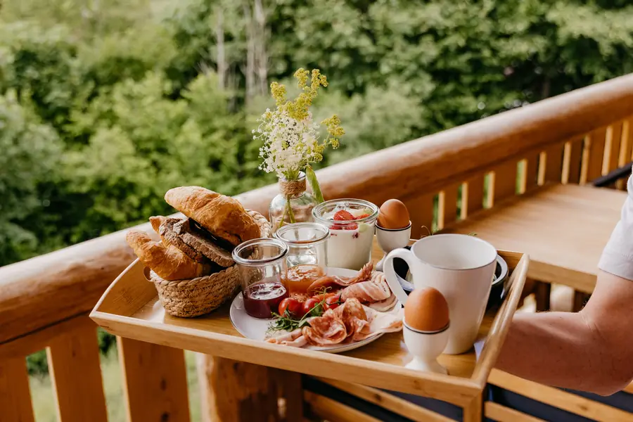 Frühstückstablett mit Brötchen, Eiern und Kaffee auf dem Balkongeländer vor dem Wald