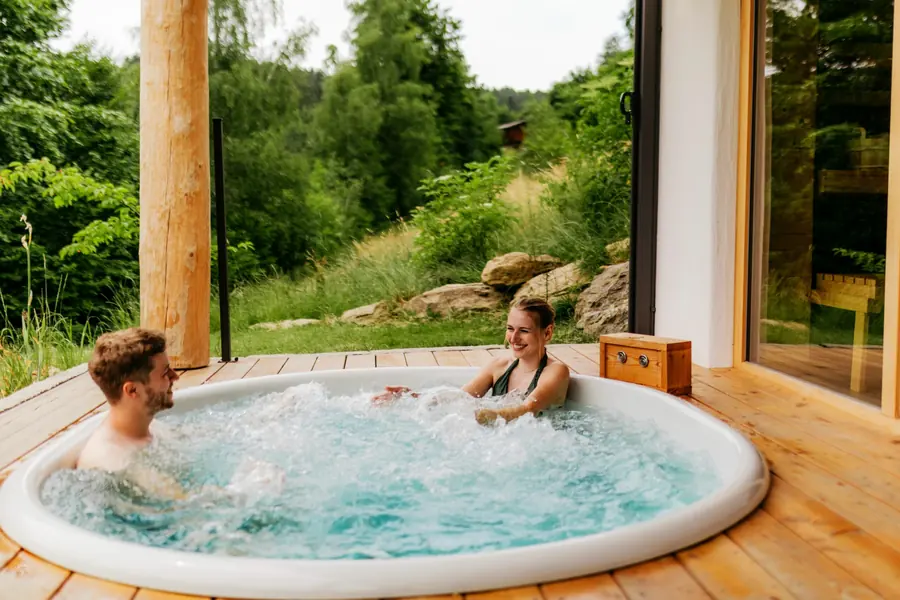 Paar im Whirlpool auf der Holzterrasse eines Chalets, dahinter Wald